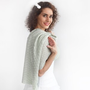 May include: A woman wearing a light green lace shawl with a white top. The shawl is draped over her shoulders and tied at the neck.