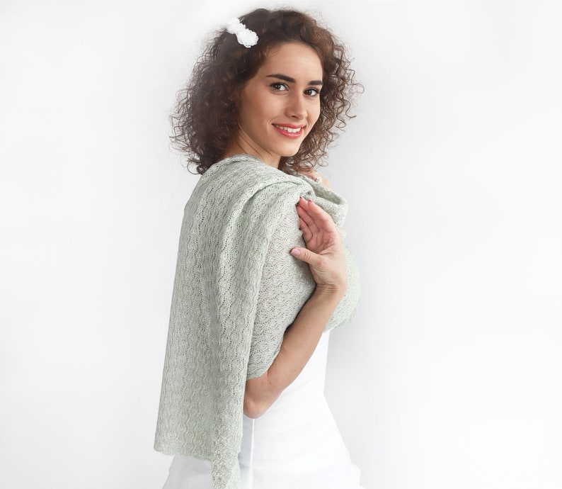 May include: A woman wearing a light green lace shawl with a white top. The shawl is draped over her shoulders and tied at the neck.