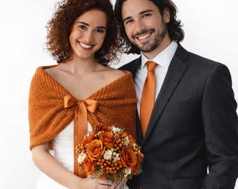 May include: A burnt orange knitted shawl with a matching satin ribbon bow, worn over a white dress, paired with a charcoal grey suit jacket, a white collared shirt, a burnt orange necktie, and a bouquet of orange roses and berries.