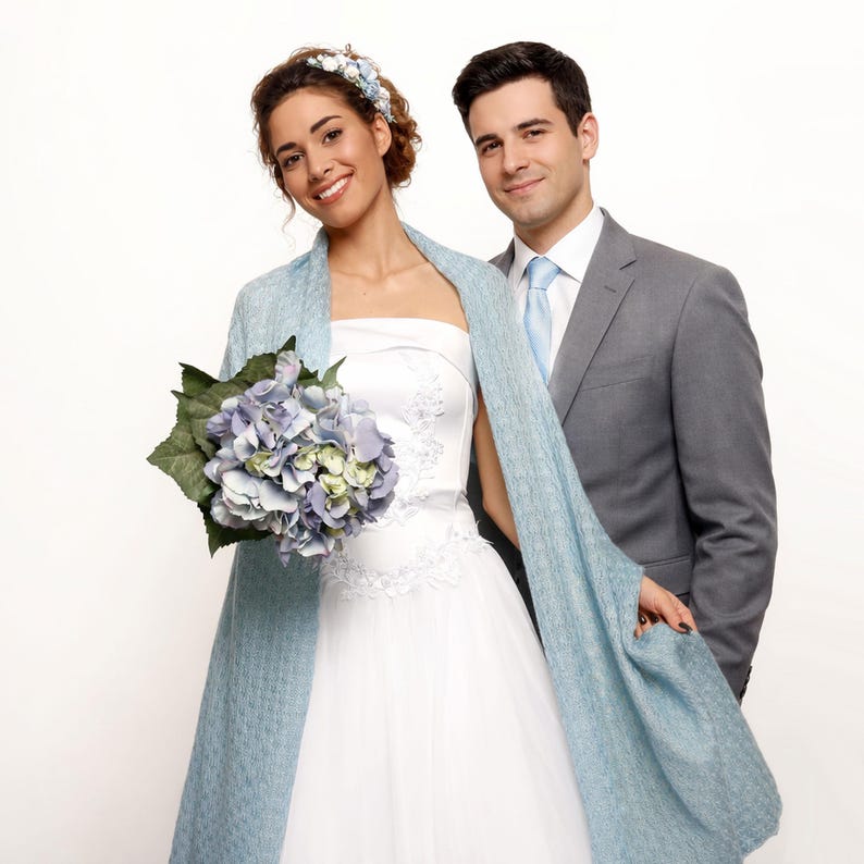 May include: A bride and groom pose for a photo. The bride wears a white strapless wedding dress and a light blue shawl. She holds a bouquet of blue and green flowers. The groom wears a gray suit with a light blue tie.