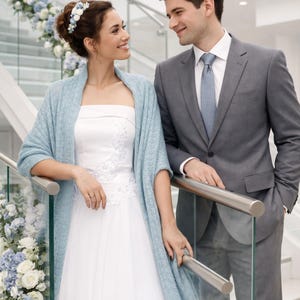 May include: A bride in a white wedding dress with floral embroidery and a light blue shawl stands next to a man in a gray suit. The background features a staircase decorated with white and blue flowers.