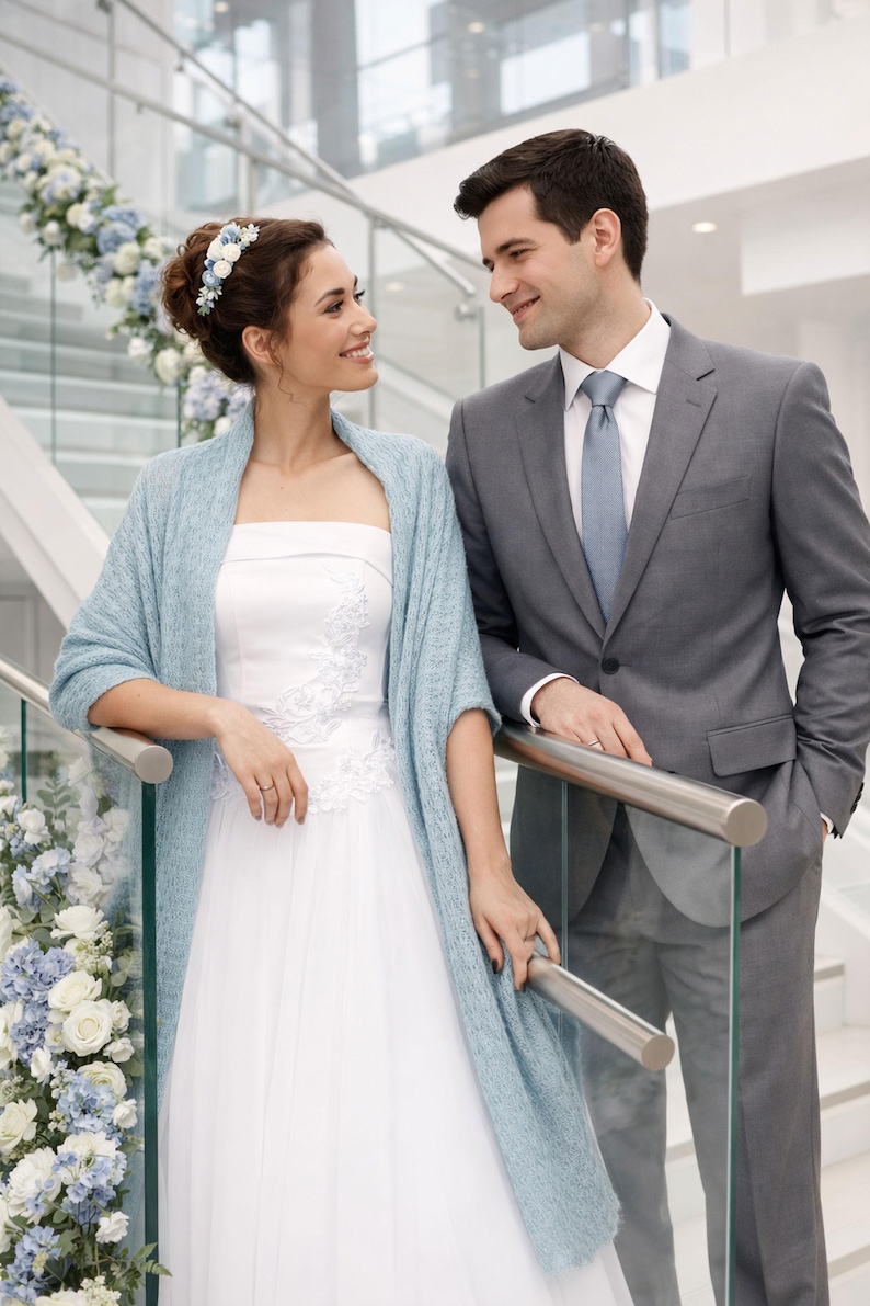 May include: A bride in a white wedding dress with floral embroidery and a light blue shawl stands next to a man in a gray suit. The background features a staircase decorated with white and blue flowers.