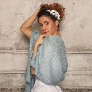 May include: A woman wearing a light blue shawl with a delicate, open-weave pattern. She is also wearing a white dress and a floral headband with blue and white flowers. The background is a textured, light gray wall.