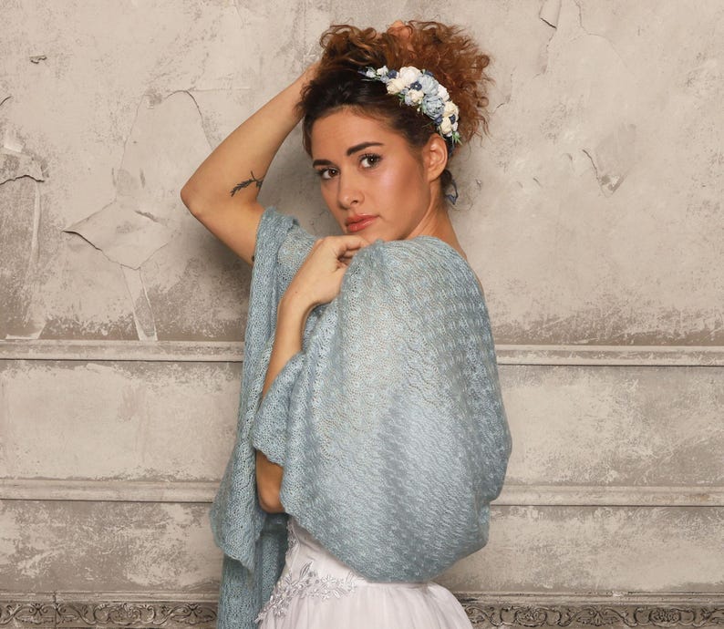 May include: A woman wearing a light blue shawl with a delicate, open-weave pattern. She is also wearing a white dress and a floral headband with blue and white flowers. The background is a textured, light gray wall.