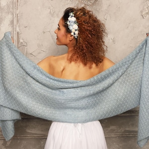 May include: A light blue shawl with a textured pattern is held open, showcasing its full length. The shawl is draped over a person wearing a white dress. A floral hairpiece with blue and white flowers adorns the person's hair.