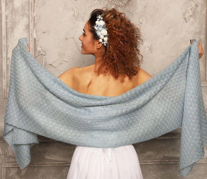 May include: A light blue shawl with a textured pattern is held open, showcasing its full length. The shawl is draped over a person wearing a white dress. A floral hairpiece with blue and white flowers adorns the person's hair.