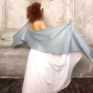 May include: A light blue woven shawl is draped over the shoulders, with a white dress visible below. The shawl has a textured pattern and is spread out, creating a flowing effect. The background is a textured wall.