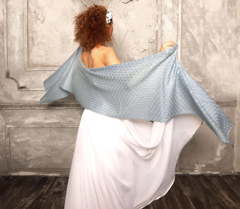 May include: A light blue woven shawl is draped over the shoulders, with a white dress visible below. The shawl has a textured pattern and is spread out, creating a flowing effect. The background is a textured wall.
