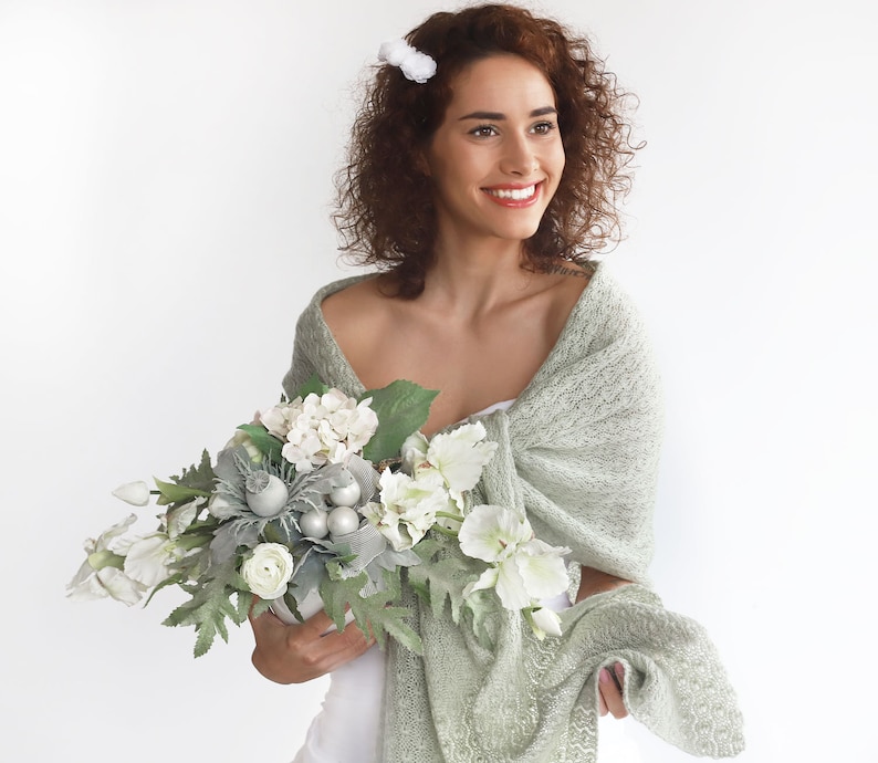 May include: A woman wearing a light green knitted shawl holds a bouquet of white and green flowers. The flowers are arranged in a white vase. The woman is smiling and looking to the side.