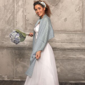 May include: A bride in a white wedding dress and a light blue shawl, holding a bouquet of blue flowers. The shawl has a textured pattern. The bride is smiling and wearing a floral headband. The background is a textured, light gray wall.