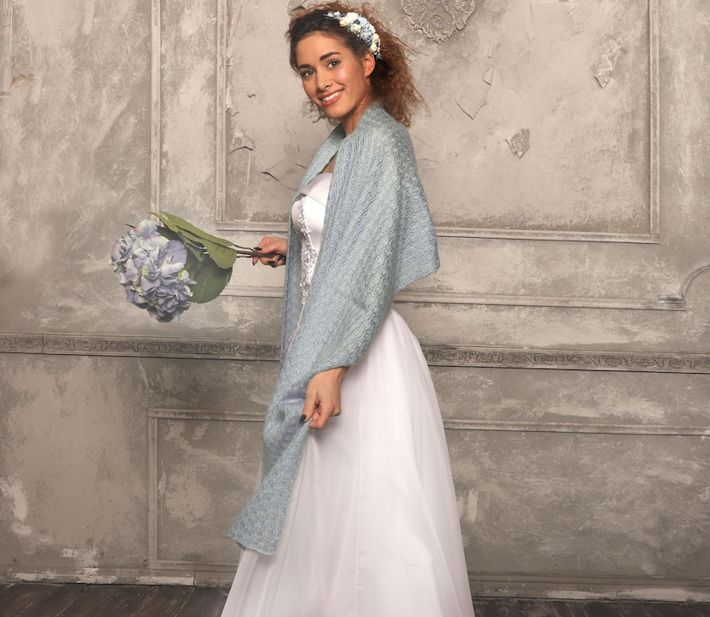 May include: A bride in a white wedding dress and a light blue shawl, holding a bouquet of blue flowers. The shawl has a textured pattern. The bride is smiling and wearing a floral headband. The background is a textured, light gray wall.