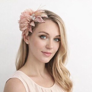 Op de afbeelding: Een blushkleurig haaraccessoire met een grote, gelaagde bloem van veren en stof. De bloem is versierd met een cluster van sprankelende strass-steentjes en delicate veertjes. Het haarstuk is ontworpen om aan de zijkant van het hoofd te worden gedragen.
