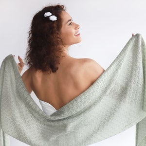 May include: A light green knitted shawl with a delicate lace pattern. The shawl is draped over a person's shoulders and back, with one end held in their hand.