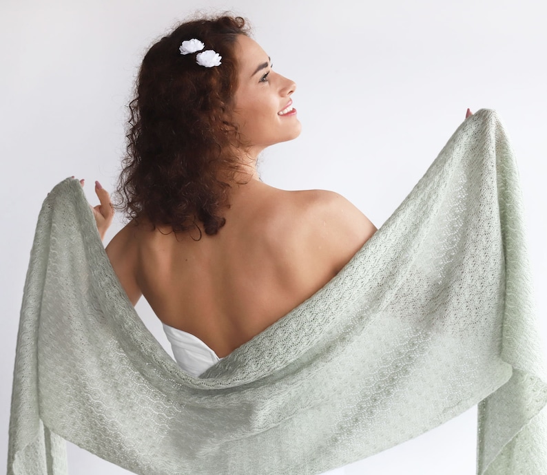 May include: A light green knitted shawl with a delicate lace pattern. The shawl is draped over a person's shoulders and back, with one end held in their hand.