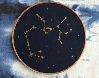 Könnte beinhalten: Ein marineblauer Stickrahmen mit einer goldenen Stickerei der Sternbilder des Schützen.