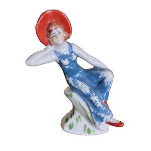 May include: A porcelain figurine of a woman in a blue dress and a red hat, reclining on a white base. The figurine has red shoes and is painted with delicate details. The base has green leaf accents.