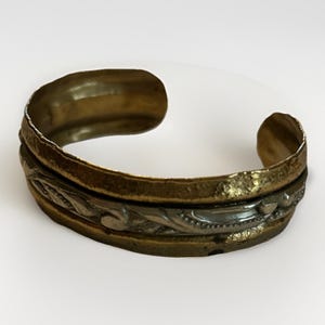 May include: A vintage-style cuff bracelet with a textured, tarnished brass finish. The bracelet features a central band with a silver-colored, ornate floral design. The open cuff design allows for easy wear. A unique piece of jewelry.