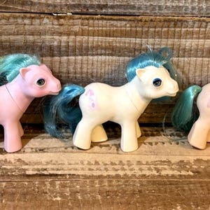 Puede incluir: Tres ponis de juguete vintage en colores pastel. Los ponis tienen crines y colas azules, y cada uno tiene un color de cuerpo diferente: rosa, blanco y melocotón. Los ponis están sobre una superficie de madera.