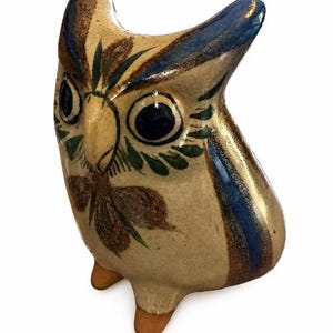 May include: A ceramic owl figurine featuring a cream-coloured base with hand-painted details in dark blue, green, and brown. The owl has large, dark circular eyes, a prominent beak, and two small, unglazed orange-brown feet at the base.
