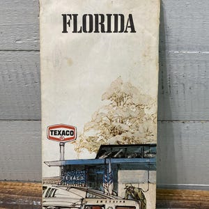 May include: Vintage Florida travel brochure with the word "FLORIDA" in bold black letters at the top. The illustration depicts a Texaco gas station, a classic car, and a person. The brochure has a worn, aged appearance.