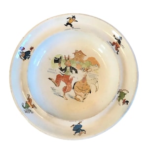 May include: A vintage white ceramic plate with a colorful illustration of cartoon characters. The central image features a rabbit in a suit and a chicken. The plate has text that reads "Uncle Wiggily" and "Jackie Bowwow".