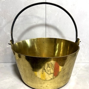 Puede incluir: Un cubo de latón antiguo con un asa curva de metal negro. El cubo tiene una base redondeada y una abertura ancha. El latón tiene una superficie pulida y reflectante. El asa está sujeta a los lados del cubo con pequeños bucles de metal.