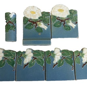 May include: A collection of blue ceramic tiles with a floral design. The tiles feature white flowers with yellow centers, green leaves, and brown stems. The tiles are arranged in a decorative pattern.