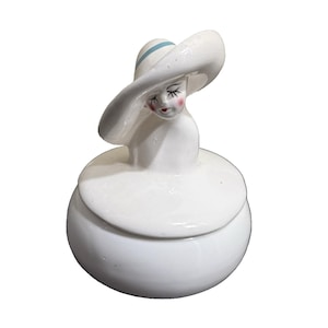 May include: A white ceramic trinket box depicting a woman in a wide-brimmed hat with a light blue band. The woman's face has closed eyes, rosy cheeks, and red lips. The box is round with a matching lid.