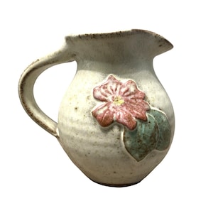 May include: Cream-colored ceramic pitcher with a handle and a spout. A raised pink and green floral design adorns the front. The pitcher has a rustic, handmade appearance, with visible texture and a speckled finish.