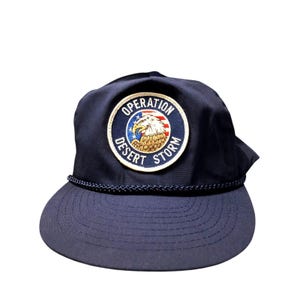 May include: A navy blue baseball cap featuring a circular embroidered patch on the front. The patch displays a bald eagle over a red, white, and blue striped background with the text OPERATION DESERT STORM. A navy blue braided cord trim sits above the brim.
