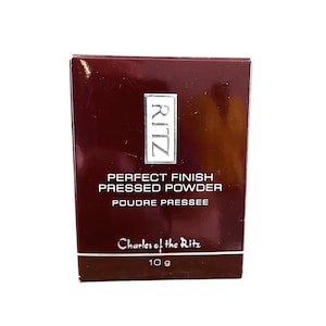 May include: A maroon box of Ritz Perfect Finish Pressed Powder. The box has the Ritz logo in a white rectangle at the top. The text "PERFECT FINISH PRESSED POWDER POUDRE PRESSEE" is printed in black. The bottom of the box reads "Charles of the Ritz" and "10 g".