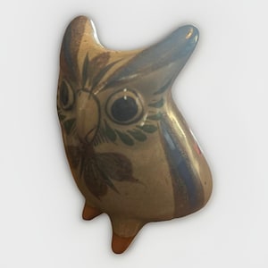 Puede incluir: Figura de búho de cerámica con base de color beige, adornada con detalles pintados en verde oscuro, marrón y azul. El búho tiene grandes ojos negros y un diseño floral estilizado en el pecho. La figura tiene un cuerpo redondeado y pies pequeños.