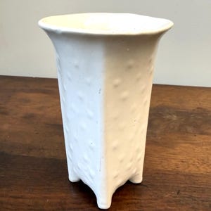 May include: A white ceramic vase with a flared rim and a square-shaped body. The exterior surface features a repeating pattern of small, raised circular bumps. The vessel is supported by four short, integrated feet at the base corners.