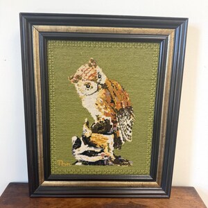 May include: A framed needlepoint embroidery featuring a brown and white owl perched on a tree stump against a textured olive green background. The artwork is enclosed in a dark brown wooden frame with a gold inner trim and includes a signature.