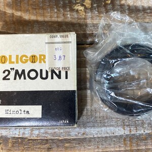 Puede incluir: Una caja vintage Soligor "T2" Mount para cámaras Minolta. La caja es crema y negra con letras doradas y una etiqueta de precio. Un adaptador de lente negro está en una bolsa de plástico transparente junto a la caja.