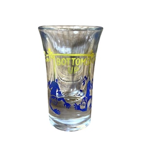 May include: Clear shot glass with the words "BOTTOMS UP" in yellow and blue monkey illustrations. The glass has a flared top and a thick base. The shot glass is empty and sits against a white background.