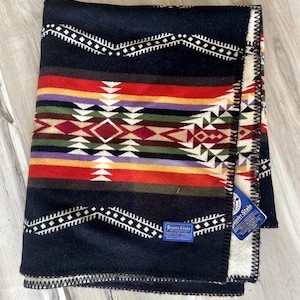 May include: A folded wool blanket featuring a black base with horizontal bands of red, orange, yellow, purple, green, and white geometric patterns. A blue Beaver State label is stitched on the front, and a blue tag is attached to the side edge.