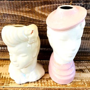 Könnte beinhalten: Zwei Vintage-Kopfvasen aus Keramik. Eine ist cremefarben mit einer Hochsteckfrisur. Die andere ist weiß mit einem rosa Hut und Schal. Beide Vasen haben die Form eines Frauenkopfes und -schultern.