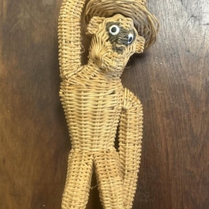 May include: A woven wicker monkey figurine in a light tan color, featuring a hat, one arm raised upward, and two round white eyes with black pupils. The figure is constructed from coiled natural fibers with a textured, hand-woven appearance.