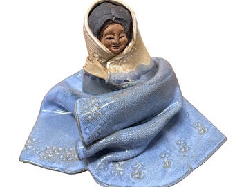 Puede incluir: Figura de cerámica de una persona envuelta en un chal y un tocado azules. La figura tiene una cara sonriente y cabello oscuro. El chal tiene un diseño estampado y ribete dorado.