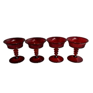 May include: Four ruby red stemmed glasses. Each glass features a round bowl with a textured pattern and a twisted stem. The base of each glass is a circular shape. The glasses are arranged in a row.
