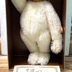 Puede incluir: Un oso de peluche blanco vintage con bozal y collar de cuero, presentado en una caja. El oso tiene una textura esponjosa y levanta el brazo. Un certificado con texto es visible en la parte inferior.