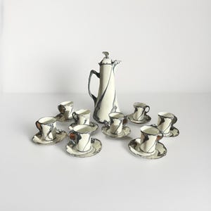 Può includere: Tazza in ceramica bianca con manico nero e la scritta "But First, Coffee" in nero. La tazza è posta su uno sfondo sfocato, che suggerisce un'atmosfera accogliente.