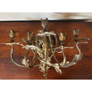 May include: A gold-toned metal wall sconce featuring four candle holders arranged on curved, leaf-shaped arms. The central mounting plate displays intricate scrollwork and foliage patterns, with a decorative finial at the top and bottom.