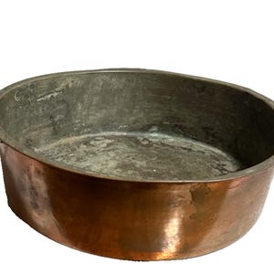May include: Oval copper pan with two handles. The pan has a dark interior and a shiny, reddish-brown exterior. The handles are square and made of the same copper material. This vintage cookware is ideal for cooking and decorative purposes.