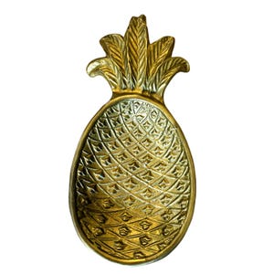 Puede incluir: Una bandeja decorativa dorada con forma de piña. La bandeja tiene una superficie texturizada detallada y una parte superior de piña estilizada. La bandeja está diseñada para guardar objetos pequeños y está hecha de metal.