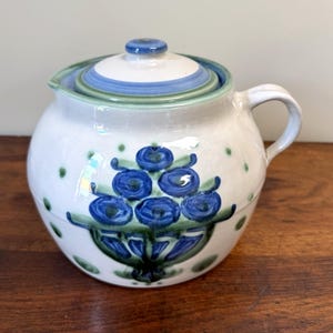 Puede incluir: Una tetera redonda de cerámica con base blanca, que presenta un motivo floral azul pintado a mano y detalles en verde. La tapa tiene anillos concéntricos azules y verdes, y el cuerpo está decorado con pequeños puntos verdes alrededor del diseño floral.