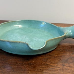 Może przedstawiać: Okrągłe, płytkie naczynie ceramiczne z jasnoturkusową glazurą, wyposażone w krótki uchwyt z jednej strony oraz mały dzióbek do nalewania na przeciwległej krawędzi. Naczynie ma gładką, lekko nierówną fakturę powierzchni i zaokrąglony brzeg.