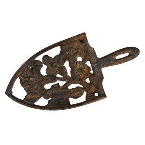 Może przedstawiać: Żeliwny podkładka pod garnek z kwiatowym wzorem. Podkładka jest w kolorze ciemnobrązowym i ma uchwyt. Ten dekoracyjny element ma chronić powierzchnie przed gorącymi naczyniami.