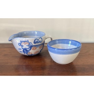 Puede incluir: Una jarra de cerámica para leche y un cuenco pequeño a juego. La jarra presenta una ilustración de un mono en azul y color melocotón sobre un fondo blanco con un borde azul. El cuenco tiene una base blanca con una banda azul en el borde.